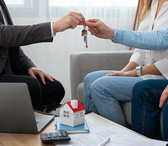 A home-finance scene: a set of keys and a small model house on a table beside a calculator, a laptop and paperwork